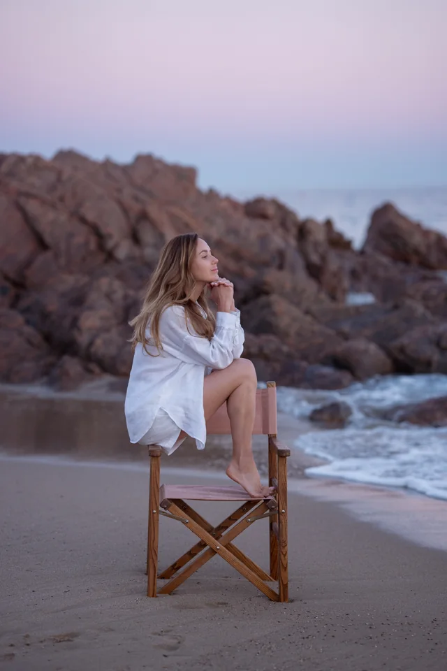 Woman in a white shirt seated on a tall chair beside rocks and the sea at dusk