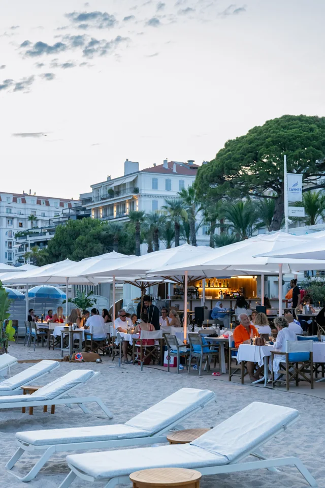 Guests dining under white parasols beside the beach, with sun loungers in the foreground
