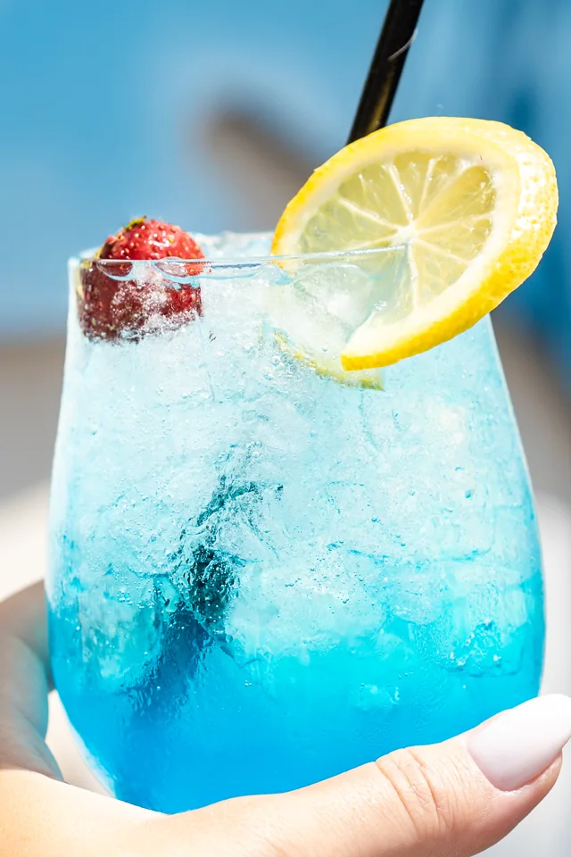 A hand holding a bright blue cocktail with ice, lemon and strawberry