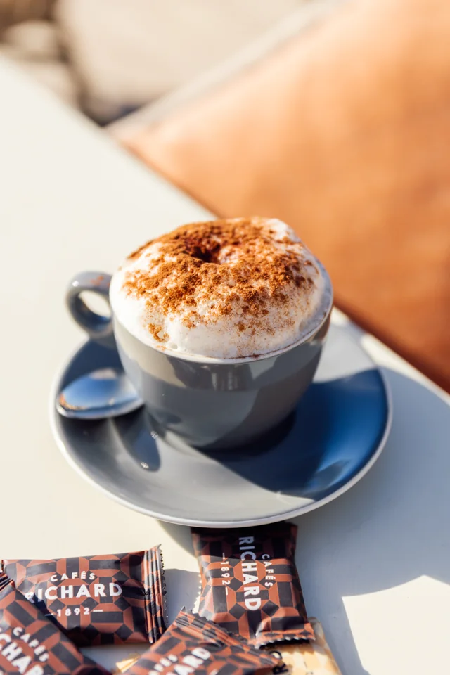 Cappuccino on a blue saucer with branded chocolate squares on a sunlit table