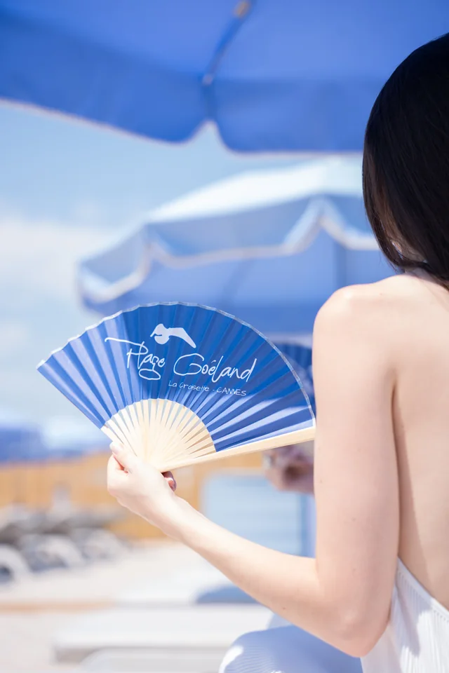 A woman holding a blue Plage Goeland fan beneath a blue beach umbrella