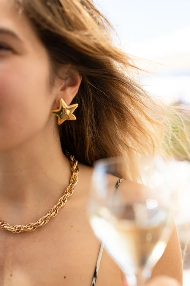Gold star earring and chain necklace with sunlit hair and a glass in the foreground